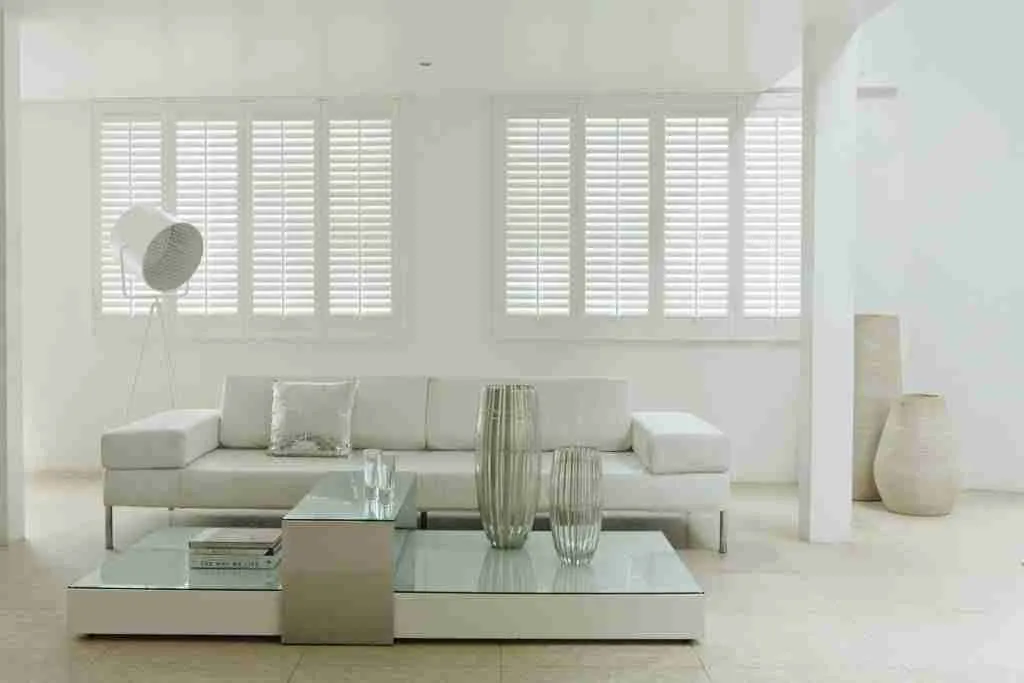 A minimalist living room decorated in white, with two large window shutters behind the sofa.